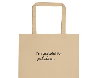 Puede incluir: Una bolsa de tela beige con el texto "I'm grateful for pilates." impreso en negro.