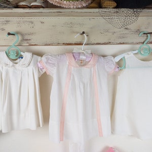 May include: Three vintage white baby dresses hanging on a wall. The dresses have different styles, including a pleated dress with a Peter Pan collar, a dress with pink ribbon accents, and a sleeveless dress. The hangers are light blue.