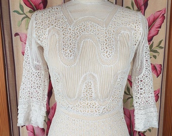 May include: A cream-colored lace dress with a high neckline and three-quarter sleeves. The dress features intricate floral and geometric patterns throughout the bodice and sleeves, with a fitted waist and a flared skirt. The dress is displayed on a mannequin.