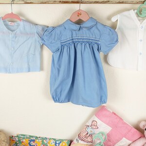 May include: Three vintage baby garments hanging on a wall. A light blue button-up shirt, a blue romper with smocking, and a white vest with a pocket. A pink quilt and a stuffed animal are in the foreground.