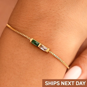 May include: A delicate gold bracelet featuring a linear design with two rectangular gemstones: one emerald green and one clear. The bracelet is worn on a wrist, showcasing the fine chain and the Brezza Jewelry logo.