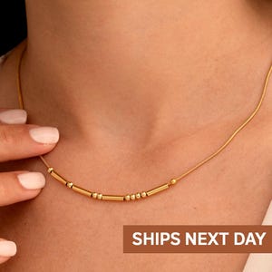 Op de afbeelding: Een delicate gouden ketting met een reeks kleine, cilindrische en bolvormige kralen. De ketting wordt om de nek gedragen. De afbeelding bevat ook de tekst "SHIPS NEXT DAY" en het merklogo "Brezza Jewelry".