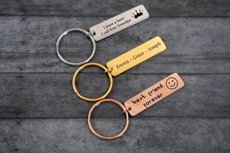 K&ouml;nnte beinhalten: Drei Schl&uuml;sselanh&auml;nger in Silber, Gold und Ros&eacute;gold mit gravierten Botschaften. Der silberne Schl&uuml;sselanh&auml;nger sagt "Ich habe einen Helden, den ich Opa nenne" mit einem Kronensymbol. Der goldene Schl&uuml;sselanh&auml;nger sagt "Emma - Grace - Joseph". Der ros&eacute;goldene Schl&uuml;sselanh&auml;nger sagt "bester Freund f&uuml;r immer" mit einem Smiley-Symbol.