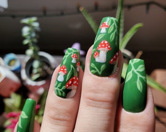 May include: A hand with long, square-shaped, painted nails. The nails are primarily green, with some featuring a design of red and white mushrooms and green leaves. One nail is solid green with a leaf design.
