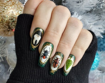 May include: A hand with long, dark green fingernails, each adorned with unique designs. One nail features a tooth, while others display insects and a leaf within ornate gold frames. The nails are set against a blurred, festive background.