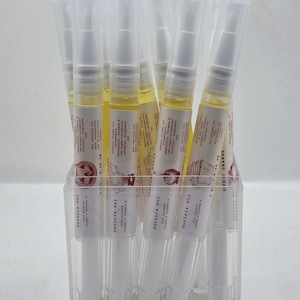 以下が含まれることがあります： 白いキャップと回転式メカニズムを備えた透明なプラスチック製のキューティクルオイルペン。黄色い液体が入っており、各ペンには黒い文字と小さな赤いイラストが描かれた白いラベルが貼られ、透明なアクリル製のホルダーに立てられています。