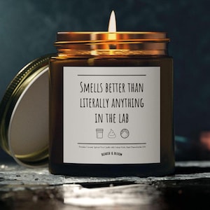 Può includere: Una candela di vetro marrone con un coperchio dorato. La candela ha un'etichetta bianca con il testo "Profuma meglio di qualsiasi cosa in laboratorio". Ci sono tre piccole icone sull'etichetta: un becher, un mucchio di cacca e una piastra di Petri.