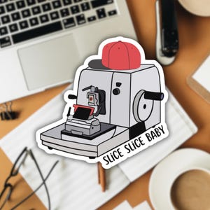May include: A white sticker featuring a cartoon illustration of a red baseball cap atop a vintage-style paper cutter. The text "Slice Slice Baby" is printed beneath the paper cutter.