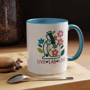 May include: White ceramic mug with a blue rim and handle. The mug features a green microscope with pink, blue, and green flowers. The text "LIVE LAB LOVE" is printed on the mug in black with blue hearts.
