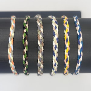 May include: Six braided bracelets in various color combinations, including blue, white, orange, green, and beige. The bracelets are displayed on a black surface.