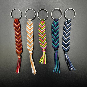 May include: Five colorful friendship bracelets with chevron patterns attached to silver key rings. The bracelets are orange, white, green, blue, and black.