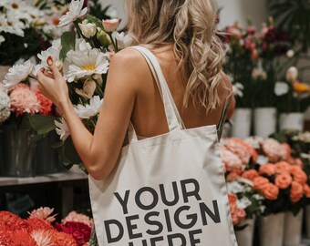 May include: A cream-colored canvas tote bag featuring the black text 'YOUR DESIGN HERE' printed on the front. The bag has two shoulder straps and is held in a setting filled with various colorful flowers in pots and vases.