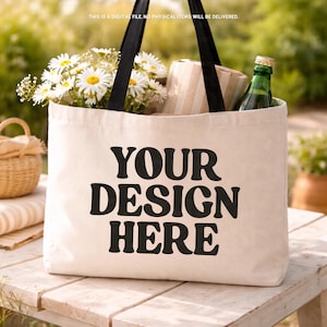 May include: A canvas tote bag with black handles and the text "YOUR DESIGN HERE" in bold black letters. The bag is filled with daisies, a wrapped gift, and a green bottle. The background is a blurred outdoor scene.