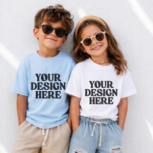 May include: Two children wearing sunglasses and t-shirts with the text "YOUR DESIGN HERE". One child wears a light blue shirt and khaki shorts, the other a white shirt and denim shorts. The background is a white wall.