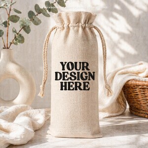 Puede incluir: Una bolsa beige para botella de vino con cordón ajustable, hecha de tela texturizada. El frente de la bolsa presenta el texto negro "YOUR DESIGN HERE" impreso en una fuente negrita y redondeada. La bolsa tiene dos cordones beige a juego.