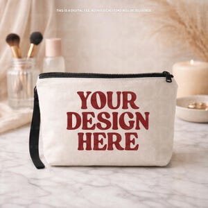 May include: A cream-colored zippered cosmetic bag with the text "YOUR DESIGN HERE" in a bold, red font. The bag has a black zipper and a black wrist strap. The background includes makeup brushes, a perfume bottle, and a candle.