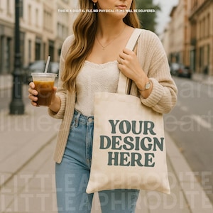 Peut inclure: Un sac fourre-tout beige avec le texte turquoise "YOUR DESIGN HERE". Le sac est porté par une personne vêtue d'un cardigan crème et d'un jean bleu. La personne tient également un gobelet en plastique transparent avec une boisson brune et une paille.