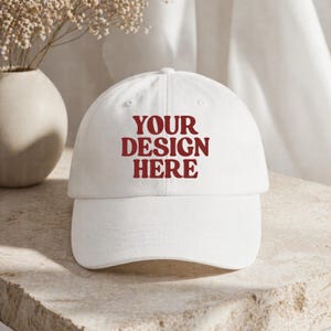 May include: A white baseball cap with a curved brim. The cap features the text "YOUR DESIGN HERE" in a bold, red font across the front. The hat is made of a soft material and is suitable for everyday wear.