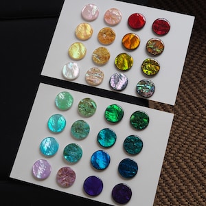 May include: A set of 24 iridescent round discs in various colours, including pink, yellow, green, blue, and purple. The discs are arranged in rows on two white surfaces.
