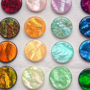 Op de afbeelding: Een verzameling kleurrijke, ronde, iriserende cabochons in verschillende tinten rood, paars, roze, oranje, groen en blauw. De cabochons hebben een glanzende, gepolijste afwerking en zijn in een rasterpatroon gerangschikt.