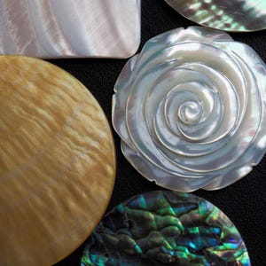 Könnte beinhalten: Nahaufnahme verschiedener irisierender Muschelteile. Ein Quadrat, ein Kreis und ein rosenförmiges Stück sind sichtbar, zusammen mit anderen Muschelfragmenten. Die Muscheln zeigen eine Reihe von Farben, darunter Weiß, Gold, Grün und Blau, mit einem perlmuttartigen Glanz.