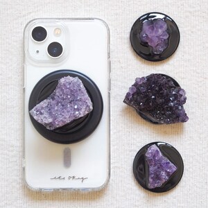 以下が含まれることがあります： 紫のアメジストクリスタルが付いた黒い電話グリップが付いた透明な電話ケース。アメジストクリスタルが付いた3つの追加の黒い電話グリップも表示されます。電話ケースには「les okay」というテキストがあります。