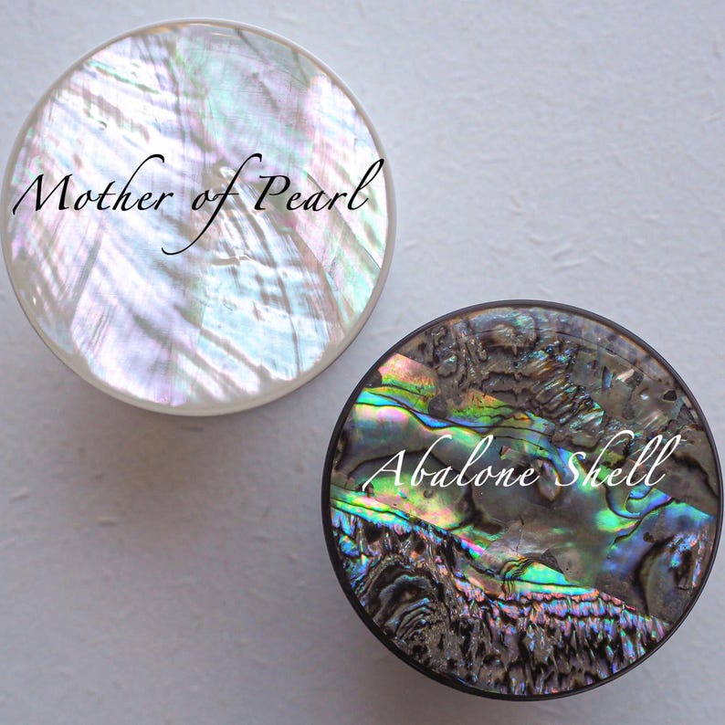 May include: Two round decorative pieces. One is iridescent white with the text "Mother of Pearl". The other is a dark, multi-colored abalone shell with the text "Abalone Shell". Both have a glossy finish.