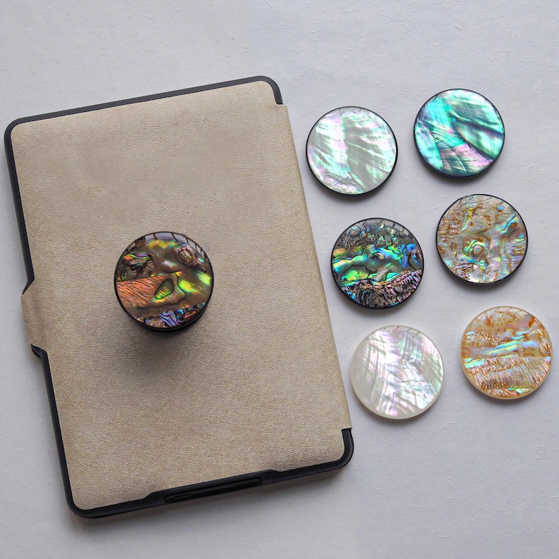 May include: A collection of iridescent abalone shell phone grips and a tablet case. The round grips feature a variety of colors, including green, blue, and gold. One grip is attached to the tan tablet case. The other grips are loose.