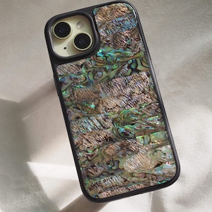 Op de afbeelding: Een zwarte smartphonehoes met een ingelegd achterpaneel van iriserende abalone-schelp. De schelp vertoont een mozaïek van glinsterende groene, blauwe en bruine tinten met een getextureerde, natuurlijke afwerking en een zwarte cameraring.