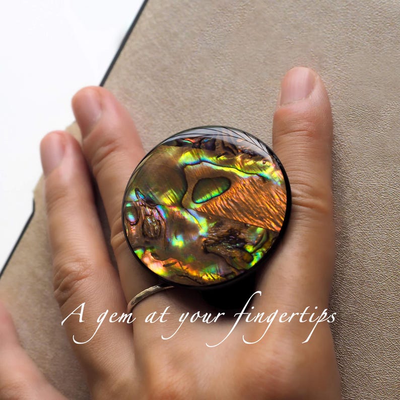 May include: A round ring with a large, iridescent abalone shell center. The shell displays a mix of green, gold, and brown hues. The ring has a black band and the text "A gem at your fingertips".