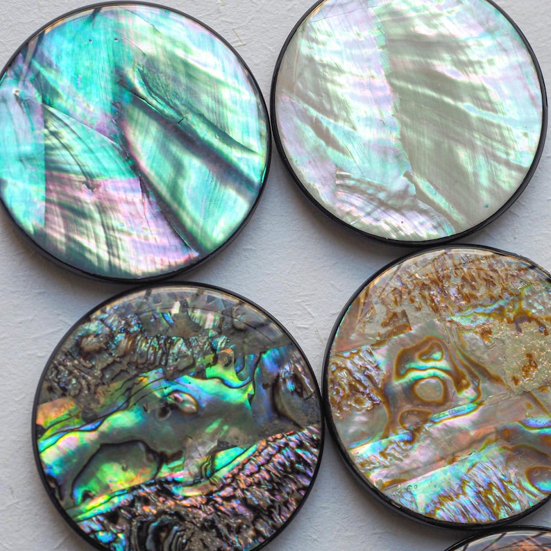 May include: Four round coasters with iridescent abalone shell surfaces. Each coaster has a black border. The abalone shells display a range of colors, including green, blue, purple, and gold, creating a shimmering effect.
