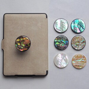 May include: A beige tablet case with a round, iridescent abalone shell phone grip attached. Six additional abalone shell grips are displayed, showcasing various colors and patterns, including green, blue, and gold.