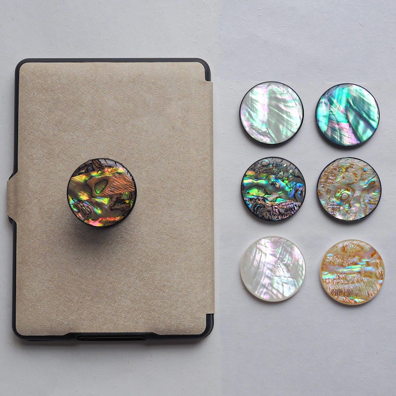 May include: A beige tablet case with a round, iridescent abalone shell phone grip attached. Six additional abalone shell grips are displayed, showcasing various colors and patterns, including green, blue, and gold.