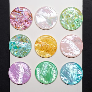 May include: A set of nine circular buttons arranged in a three-by-three grid on a white rectangular card. Each button features a distinct iridescent finish with colors including pink, white, green, yellow, purple, and blue with a pearlescent sheen.