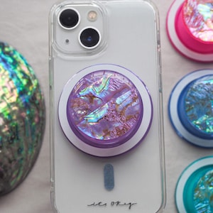 May include: A clear phone case with a purple and iridescent phone grip attached. The grip has a circular design with a pearlescent finish. Additional phone grips in pink, blue, and teal are visible. An abalone shell is in the background. The words "it's okay" are written on the phone.