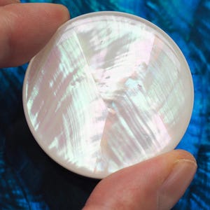 May include: A round, iridescent mother-of-pearl button. The button has a pearlescent sheen with a geometric pattern. The button is held between two fingers against a blue background.