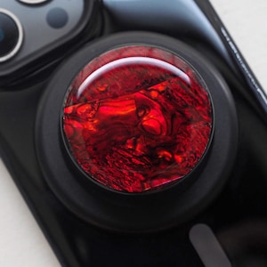 May include: A black phone grip with a vibrant red, marbled design. The circular grip features a glossy, textured surface, creating a striking contrast against the black phone case. The phone accessory is designed to enhance grip and usability.