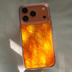 May include: Clear phone case with a vibrant orange and yellow abstract design. The case has a clear border and a camera cutout with multiple lenses. The bottom right corner features the handwritten text "ec okig".