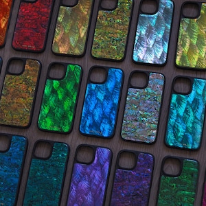 Peut inclure: Une collection d'étuis de téléphone irisés de différentes couleurs, notamment rouge, vert, bleu et violet. Chaque étui a un bord noir et un motif texturé unique, créant un effet chatoyant. Les étuis sont disposés sur une surface en bois.