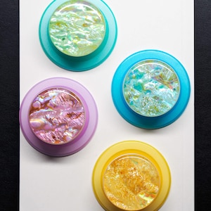 May include: Four circular phone grips featuring iridescent, shell-like inlays with swirling patterns. The grips have colorful plastic bases in teal, bright blue, light purple, and yellow, arranged on a plain white rectangular surface.
