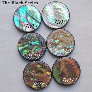May include: Six round, iridescent abalone shell cabochons in the Black Series. Each cabochon has a unique pattern of colors, including blues, greens, and browns. The text "The Black Series" is at the top.