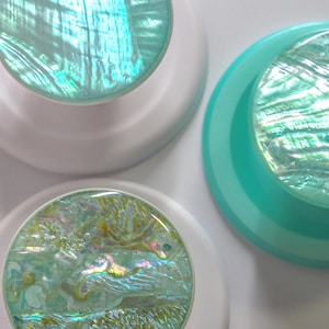 May include: Three circular knobs featuring iridescent, shell-like inlays with swirling patterns of teal, green, and pearlescent white. Two knobs have solid white bases, while one knob features a solid teal base, all arranged in a cluster.