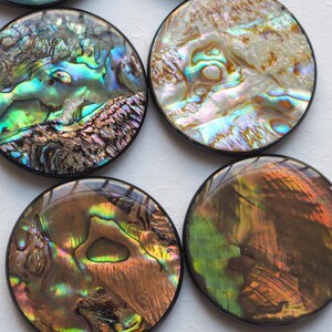 May include: Four round coasters with iridescent abalone shell inlays. Each coaster has a black border and displays a unique pattern of blues, greens, browns, and golds. The coasters are arranged in a square formation.