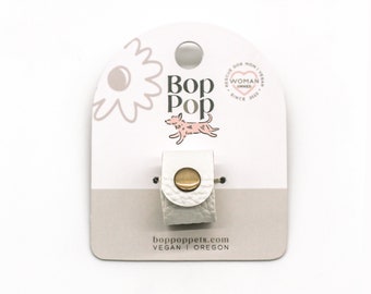 May include: A white leather dog leash holder with a gold-colored snap closure. The product is displayed on a white and gray card with the text "Bop Pop" and a pink dog illustration. The card also includes the website address boppopets.com.
