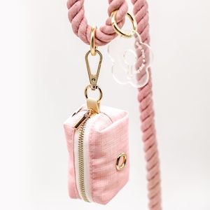 May include: A pink dog waste bag dispenser with a gold zipper and a gold ring. The dispenser is attached to a pink rope lead with a clear plastic flower charm that says "dog poop".
