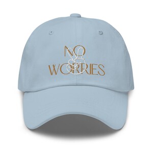 Pode incluir: Um boné de beisebol azul claro com o texto "NO WORRIES" bordado em marrom com um sinal de paz branco.