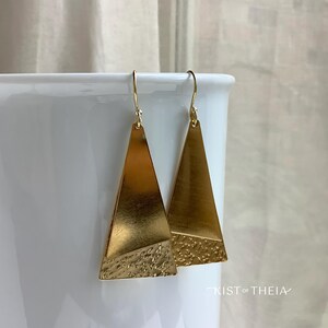 May include: A pair of gold-toned dangle earrings featuring a triangular shape with a smooth upper section and a textured, hammered lower section. The earrings hang from thin gold-toned ear wires. The text KIST OF THEIA is visible at the bottom.