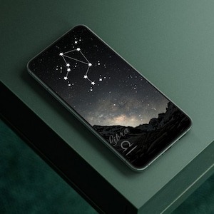 May include: A smartphone displaying a Libra constellation design against a dark night sky background. The phone rests on a dark green surface, with the word "Libra" and the zodiac symbol visible on the screen.
