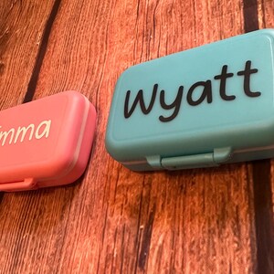 May include: Two small plastic containers with lids. One is pink with the name "Emma" in white lettering. The other is blue with the name "Wyatt" in black lettering.