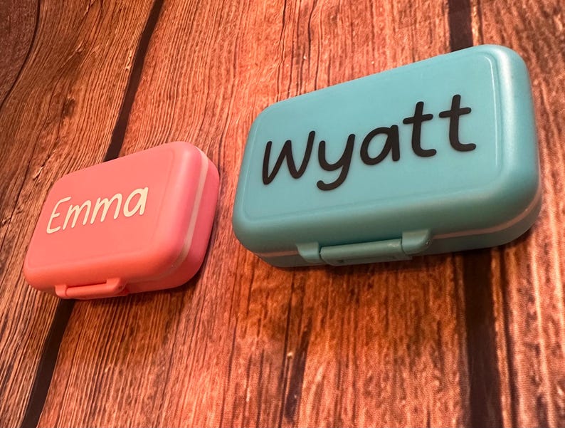 May include: Two small plastic containers with lids. One is pink with the name "Emma" in white lettering. The other is blue with the name "Wyatt" in black lettering.